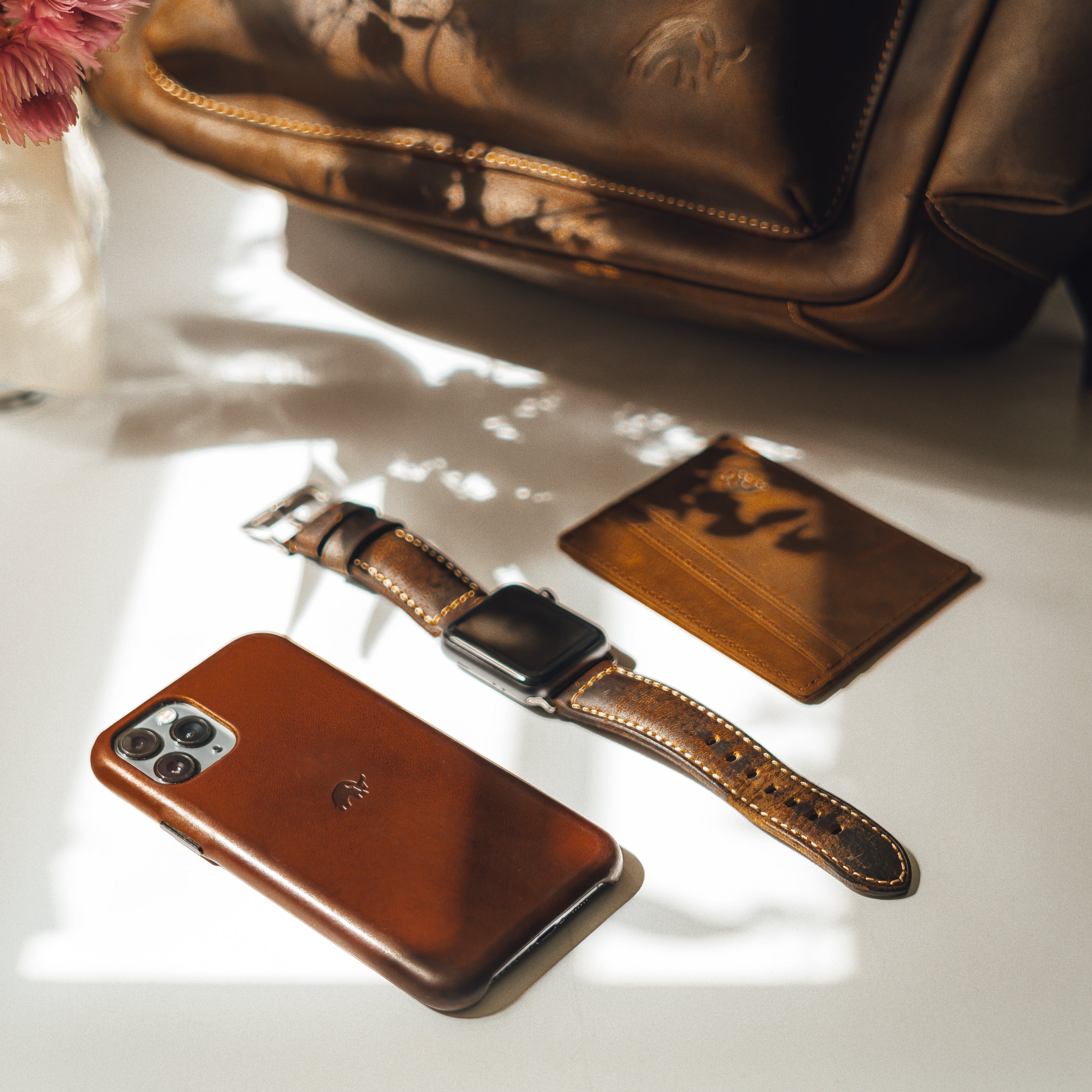 A leather card holder, a smartphone case, and a watch on a table with a floral background.