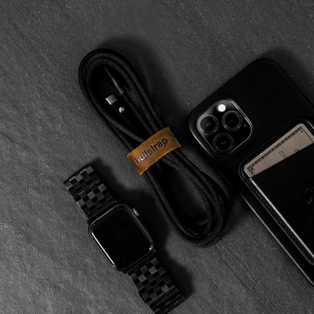 A black charging cable, an iPhone, and a black watch on a dark surface.