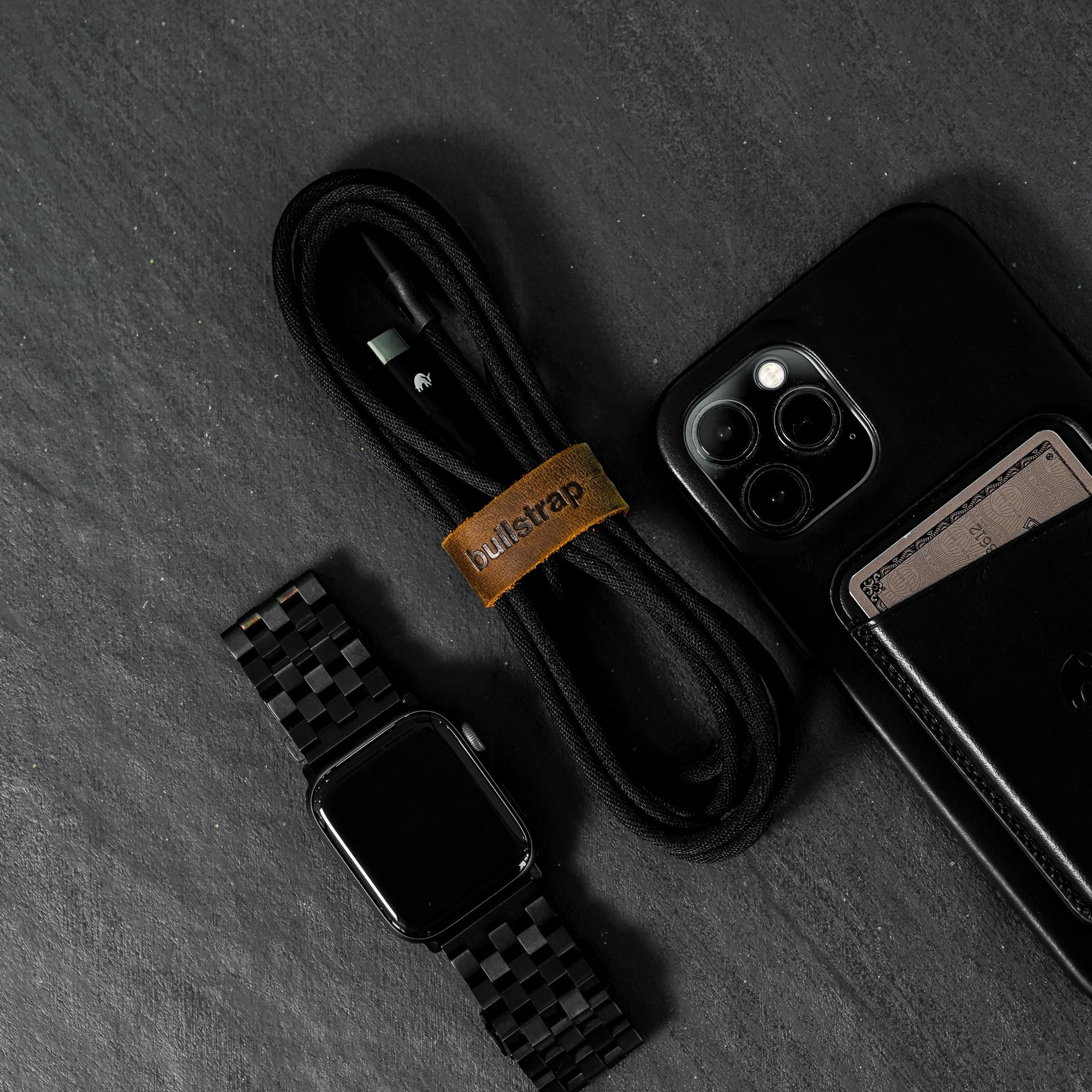 A black charging cable, an iPhone, and a black watch on a dark surface.
