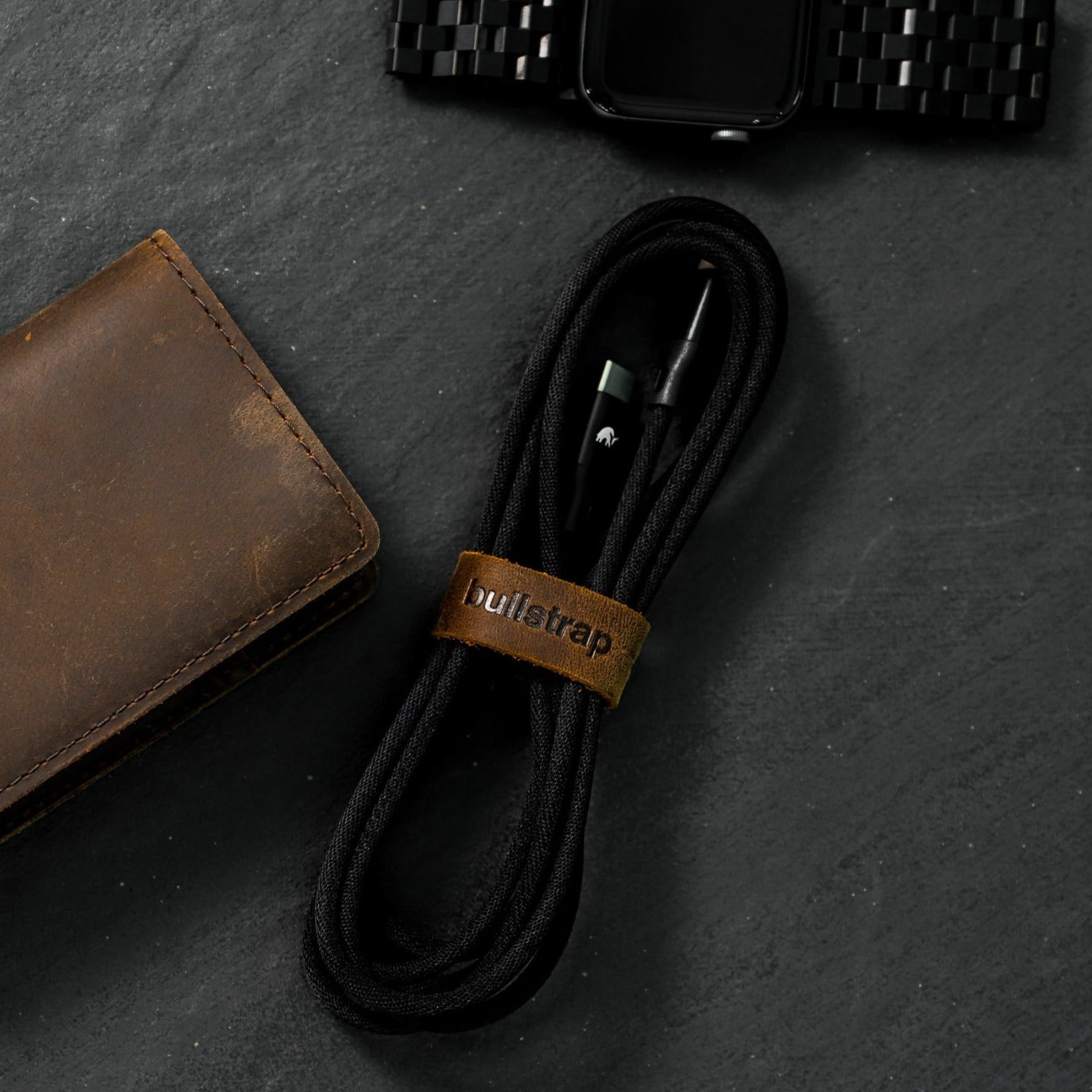 Coiled black braided charging cable secured with leather strap embossed 'bullstrap' beside brown leather wallet.