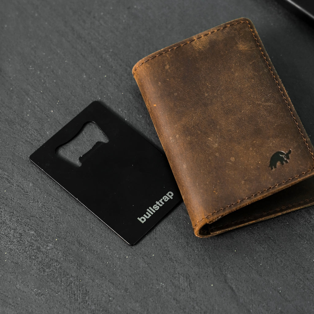 Black credit-card bottle opener stamped 'bullstrap' next to a brown distressed leather wallet with embossed mammoth logo.