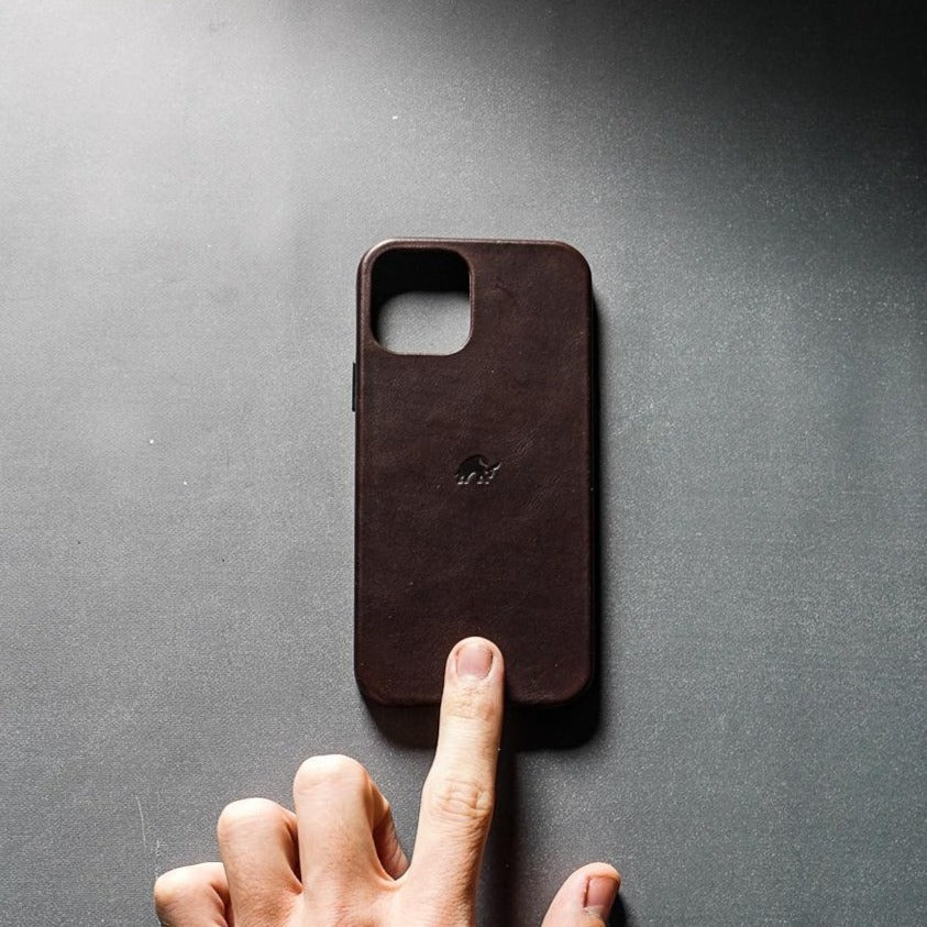 Bourbon brown leather iPhone case with small embossed elephant logo, finger touching bottom center on dark tabletop