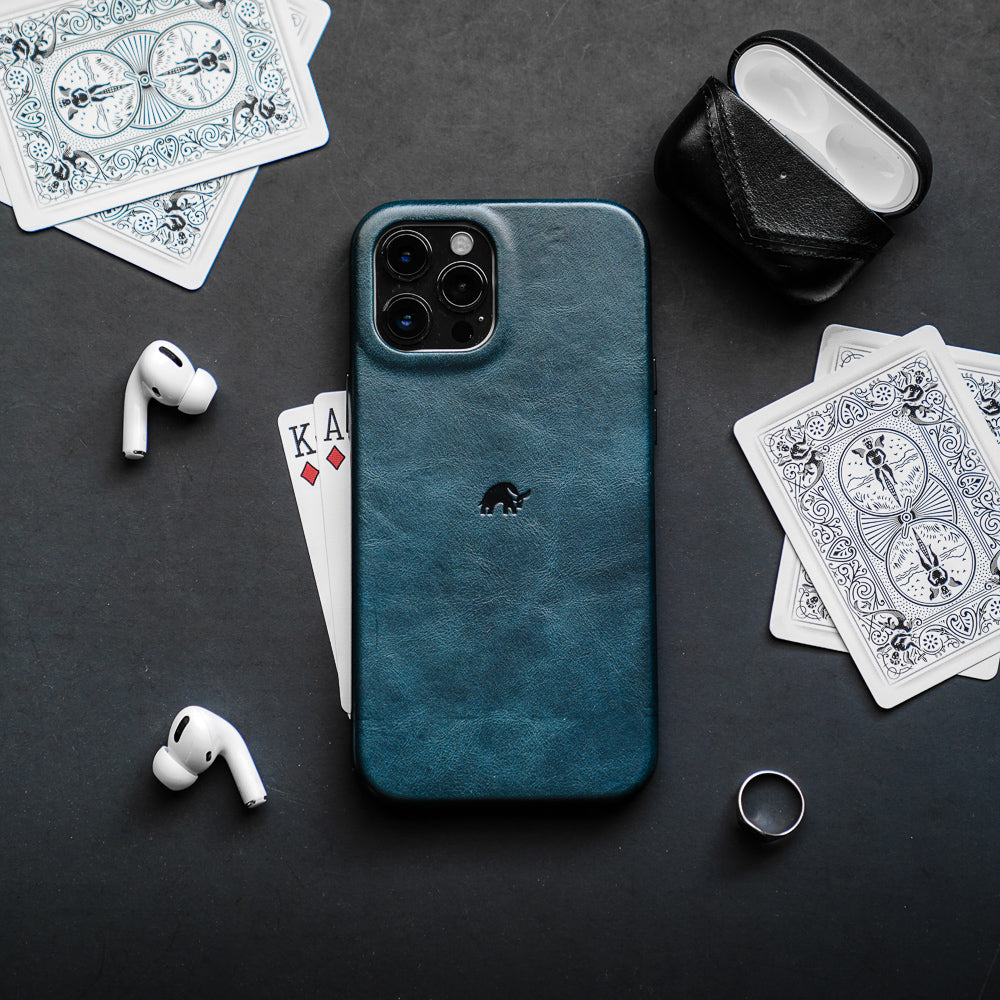 Teal leather iPhone case on dark table with playing cards showing K and A, AirPods Pro earbuds and a silver ring