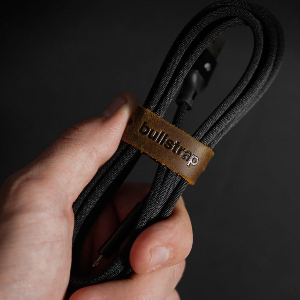 A hand holding a black charging cable with a leather strap labeled 'bullstrap'.