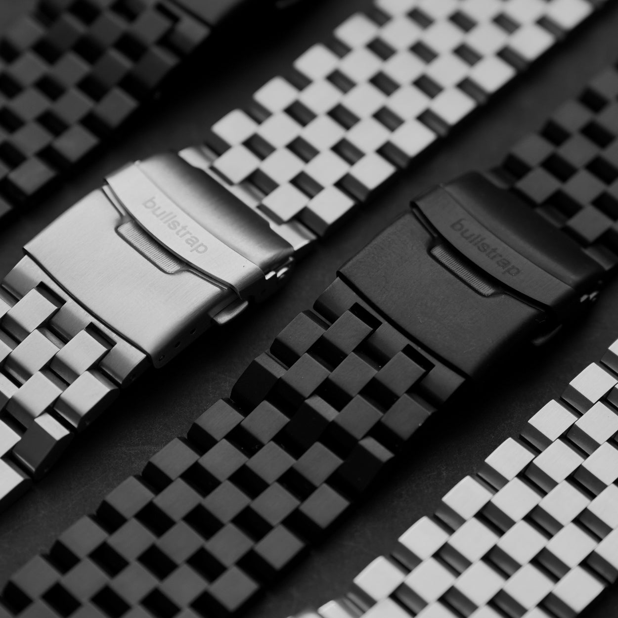Close-up of silver and black metal Apple Watch straps with linked bracelets and clasps engraved 'bullstrap'.