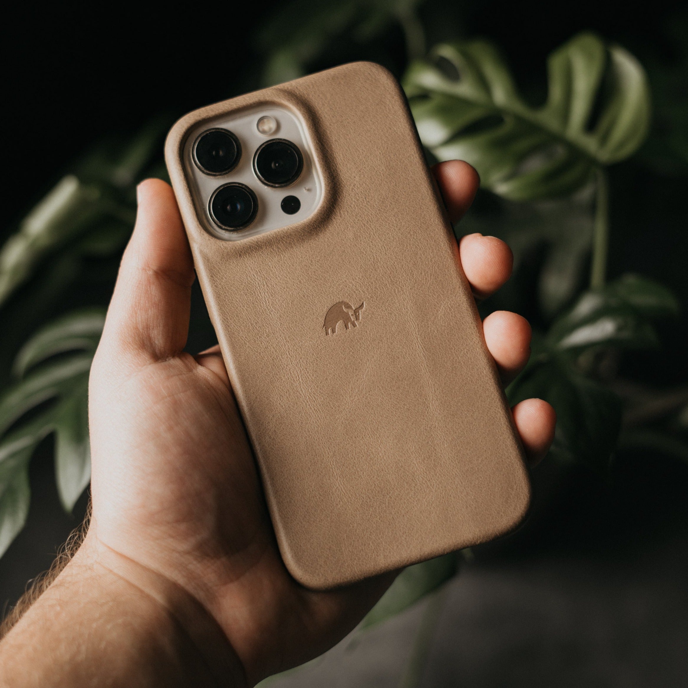 Hand holding tan leather smartphone case with triple-camera cutout and small embossed elephant logo, blurred plant background.