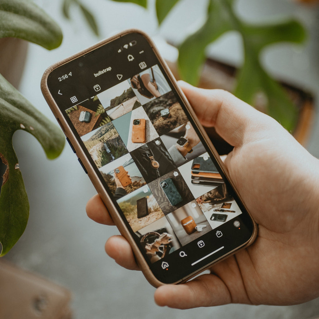 A hand holding a phone displaying various Bullstrap products including the Minimalist Portfolio Case - DUNE.