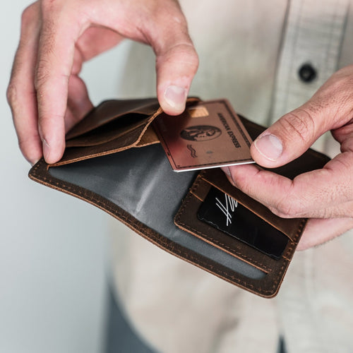 Brown leather bi-fold wallet open in hands, card slots visible and a person inserting an American Express card.