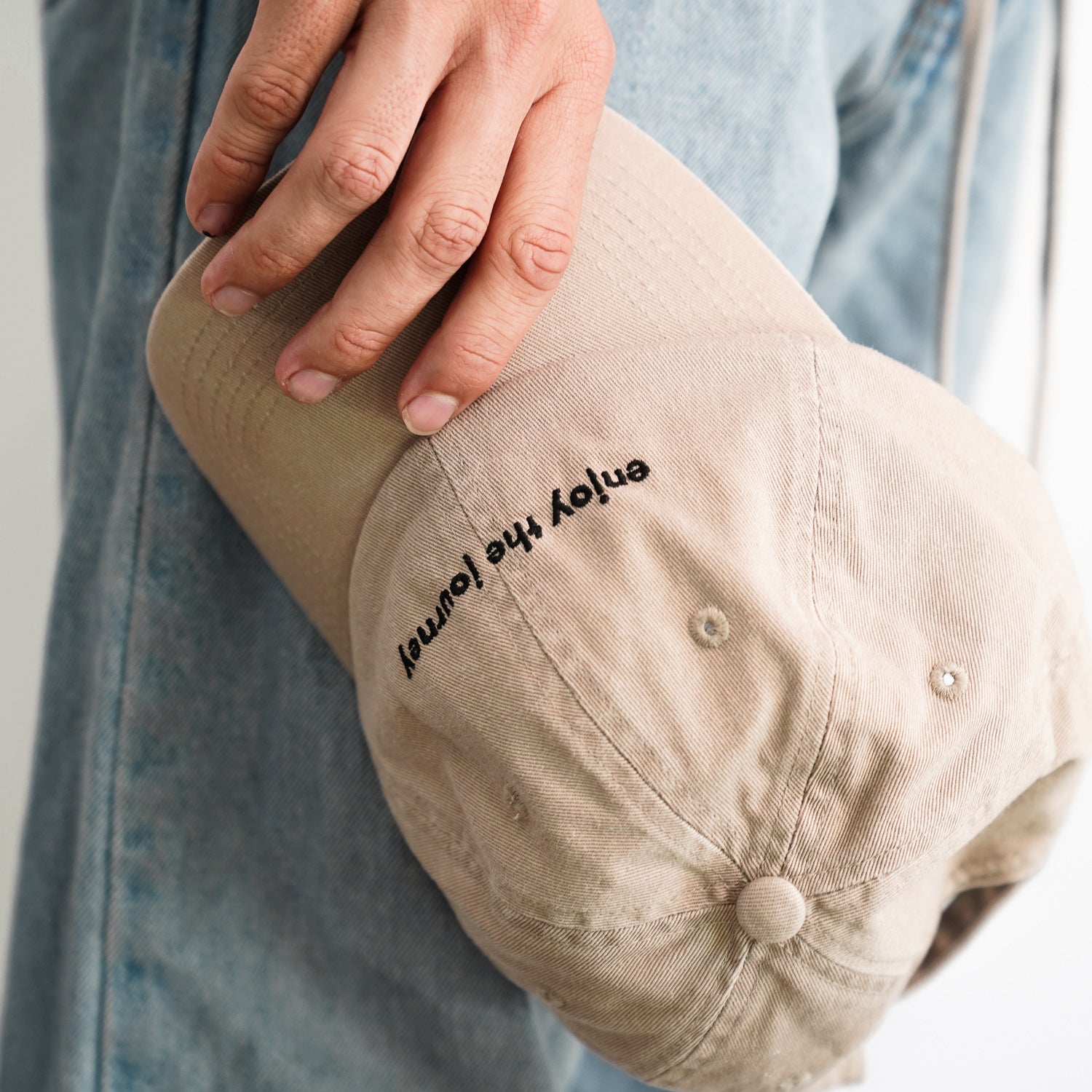 A person wearing jeans and holding an Enjoy the Journey Dad Hat in tan