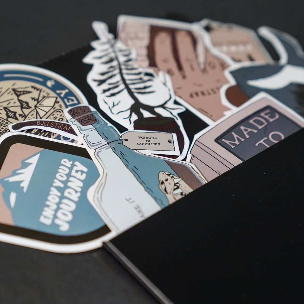 Colorful travel stickers in a black notebook. Visible text: ENJOY YOUR JOURNEY; MADE TO; DISTILLED IN FLORIDA; BULLSTRAP