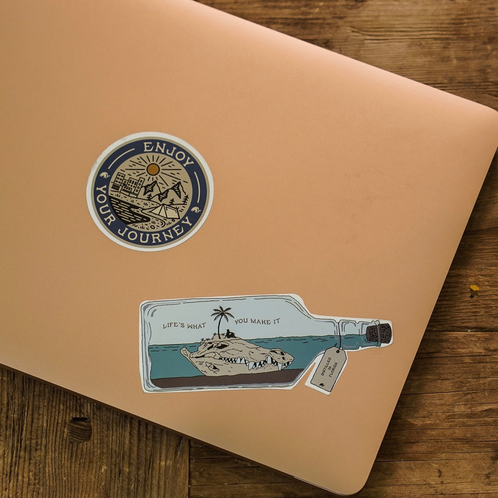 A laptop with stickers including a bottle and a circular design that says 'Enjoy Your Journey'