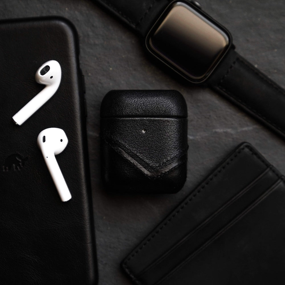 Black leather AirPods case next to AirPods, a phone, and a watch on a dark surface.