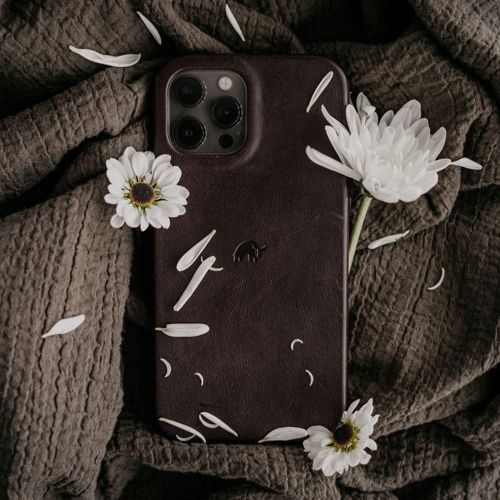 Bourbon brown leather iPhone case with embossed elephant logo on textured fabric, white daisies and scattered petals.