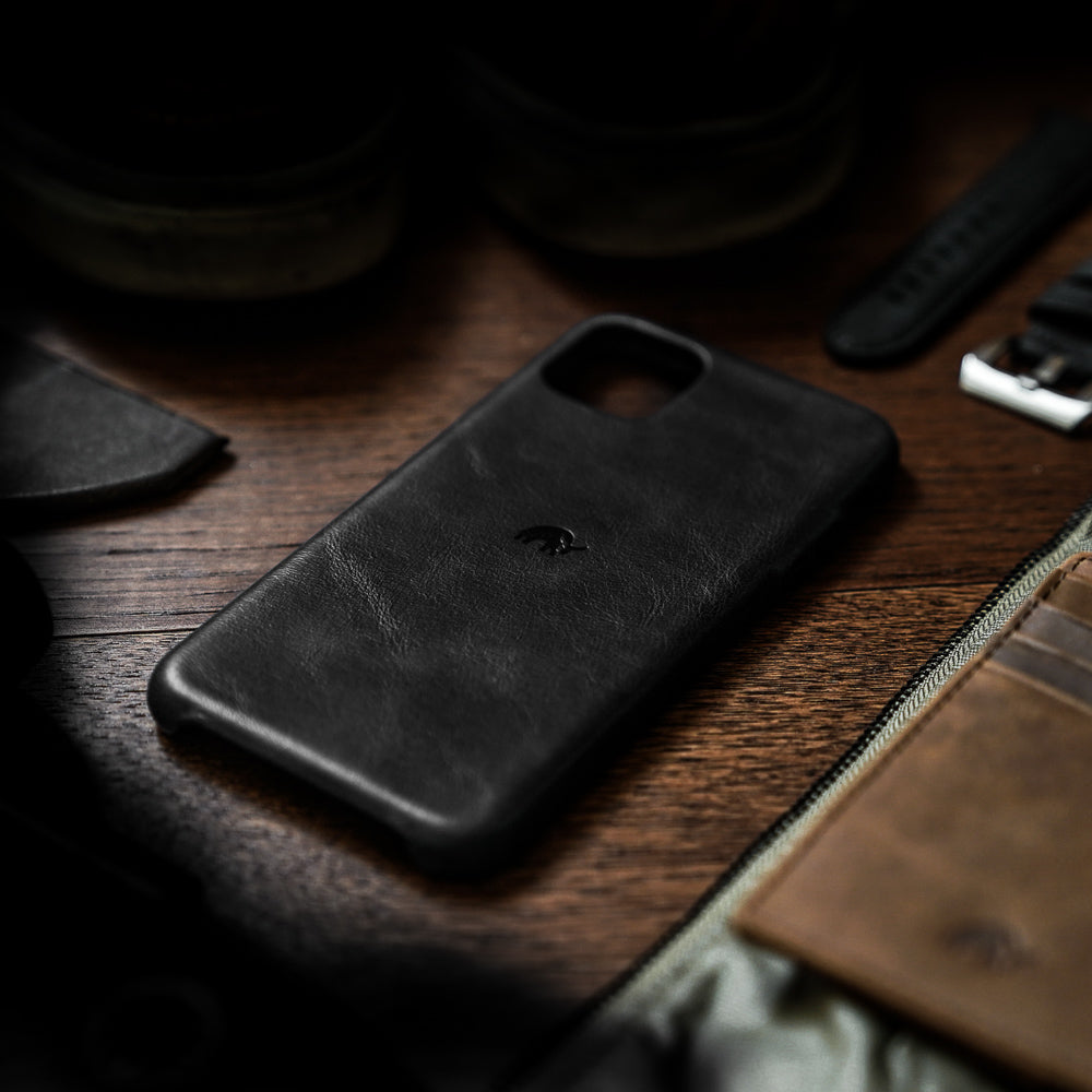 Black leather iPhone case with embossed logo on wooden table, next to a wallet and watch strap