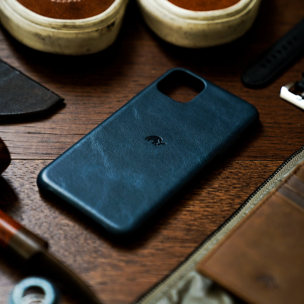 Dark blue leather phone case with small embossed animal logo on wooden table surrounded by accessories