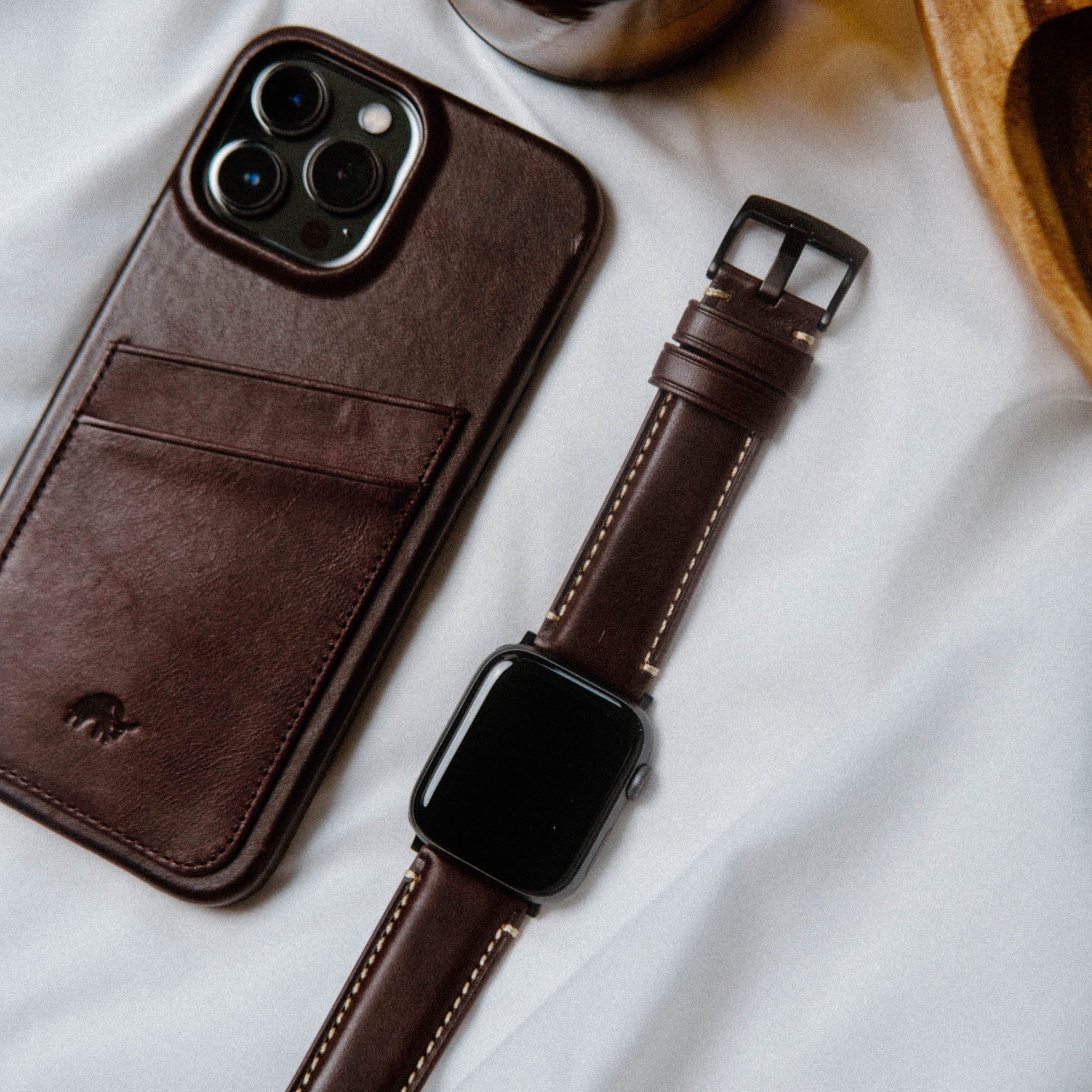 A leather Apple Watch strap in bourbon color next to a phone case on a white surface.