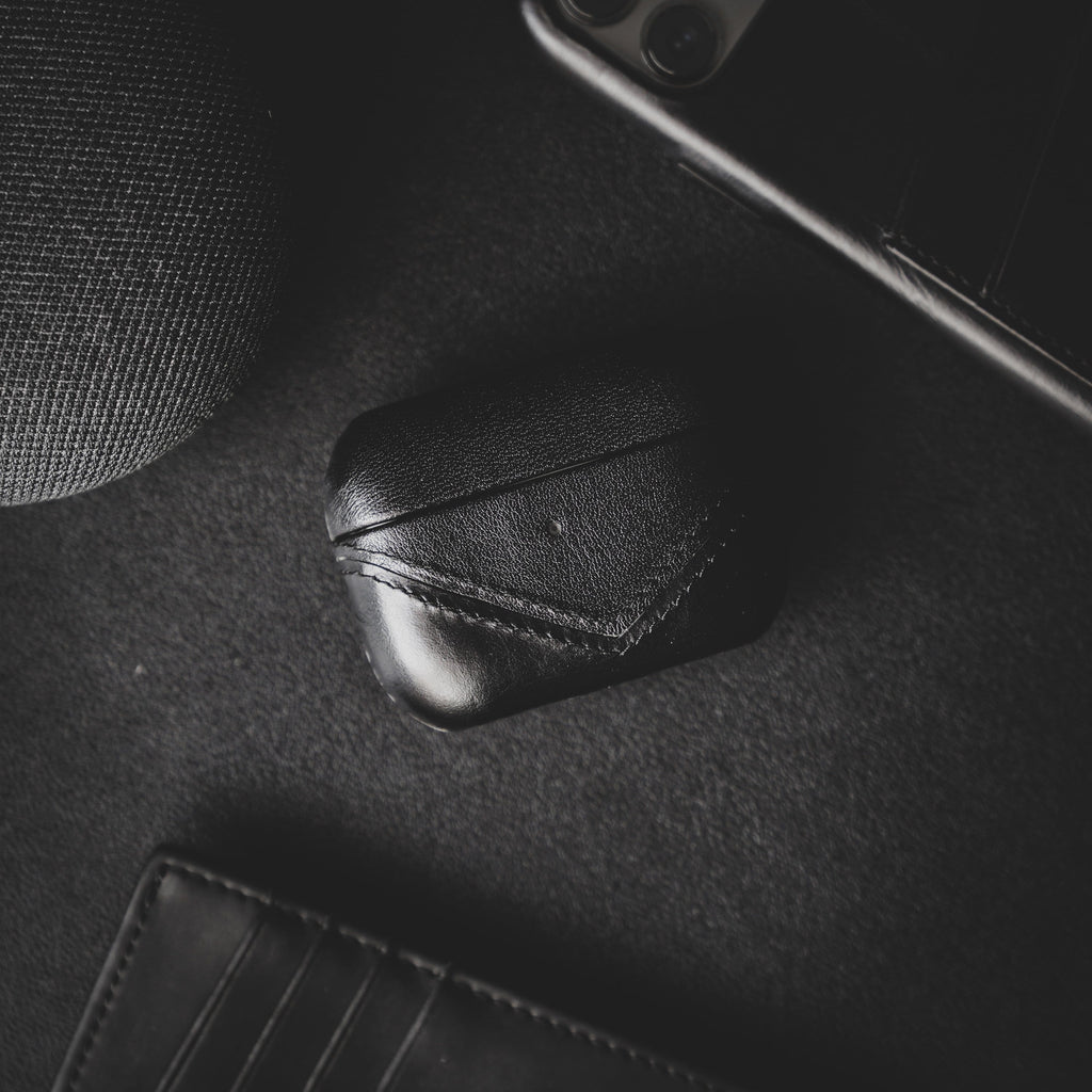 Black leather AirPods case on a dark surface with a wallet and speaker nearby