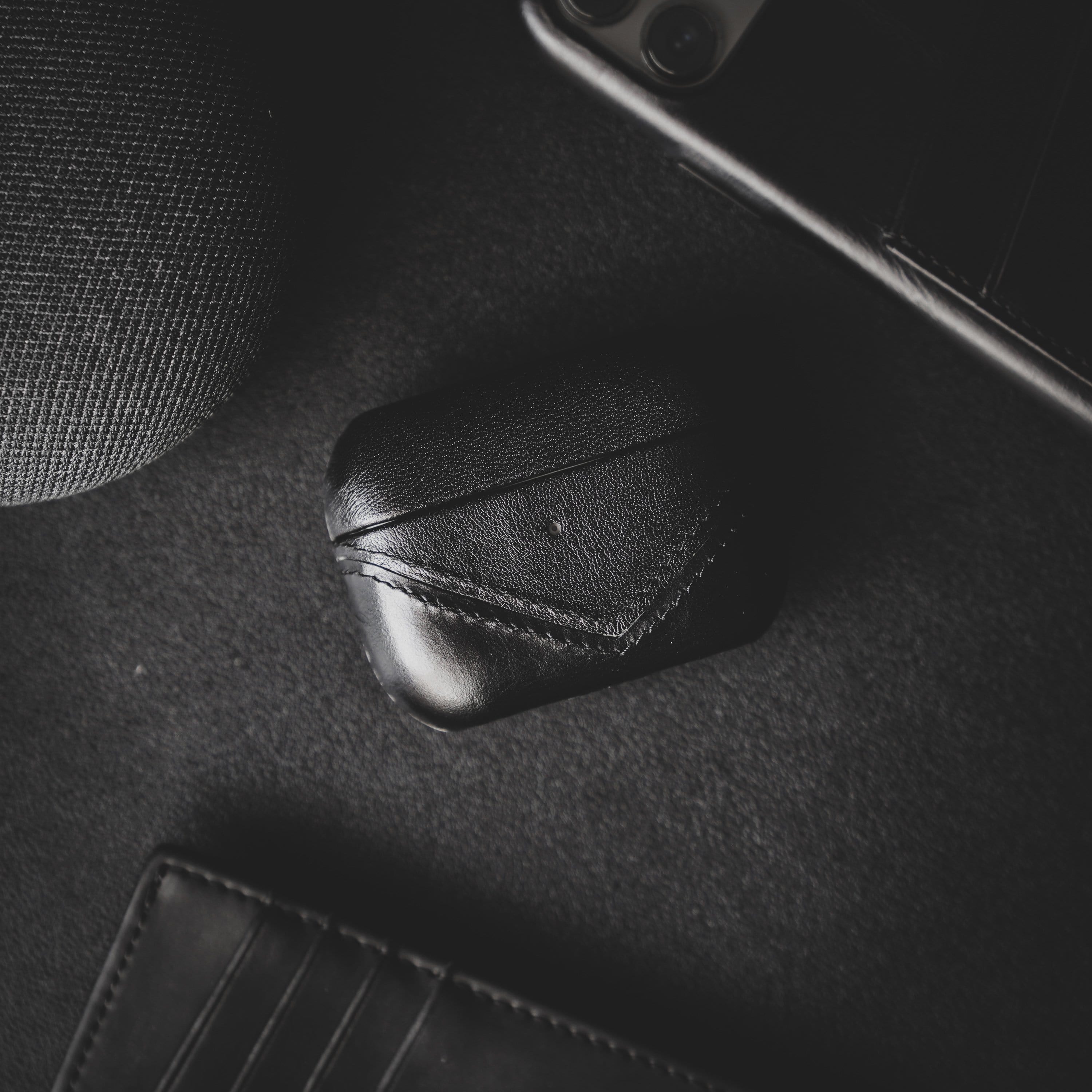 Black leather AirPods case on a dark surface with a wallet and speaker nearby