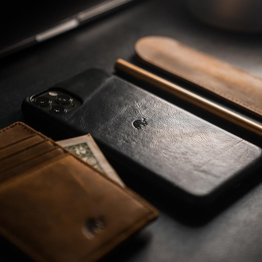 Black leather iPhone case with embossed logo beside a brown leather wallet and pen on a dark surface