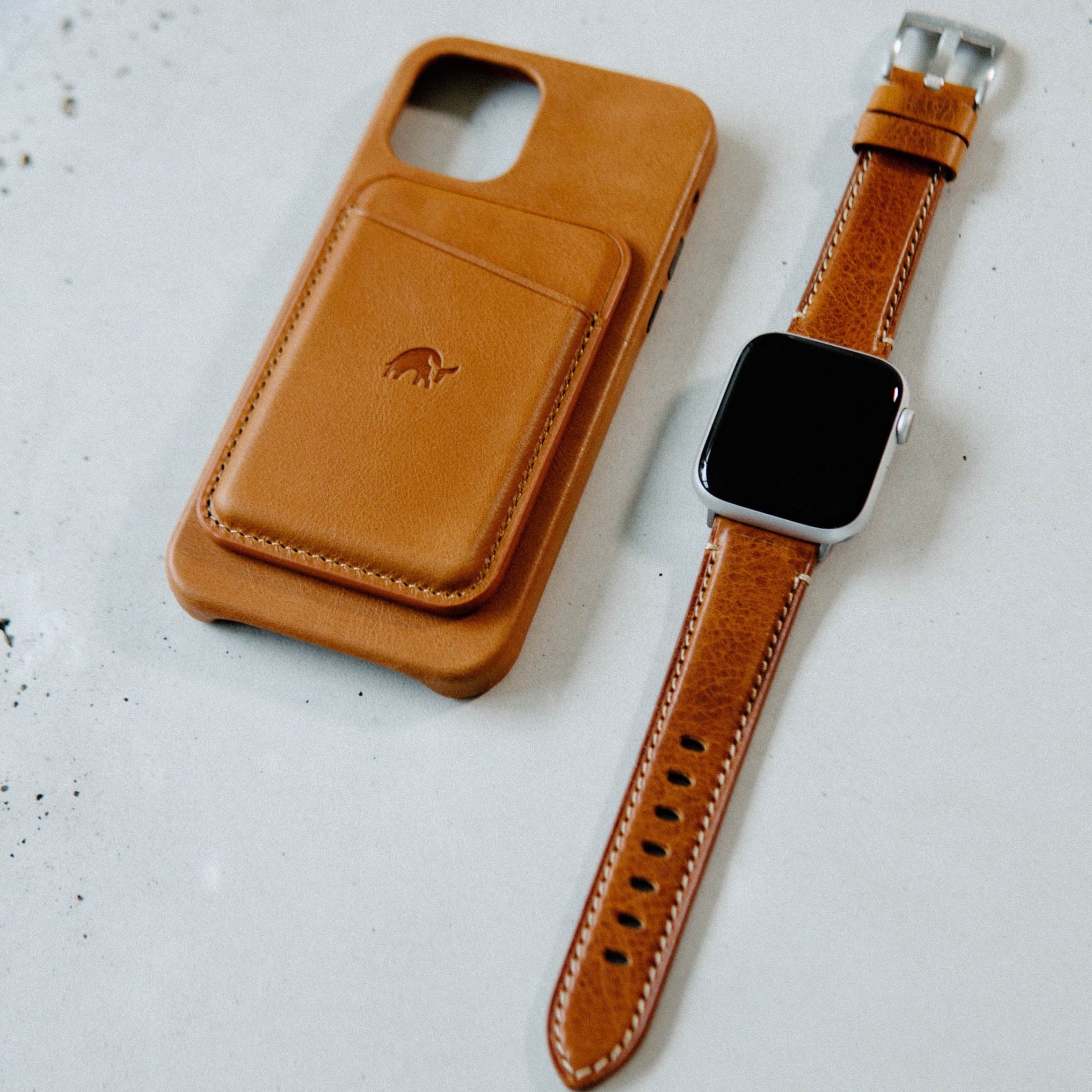 Tan leather iPhone case with card slot and brown leather Apple Watch strap on a white surface