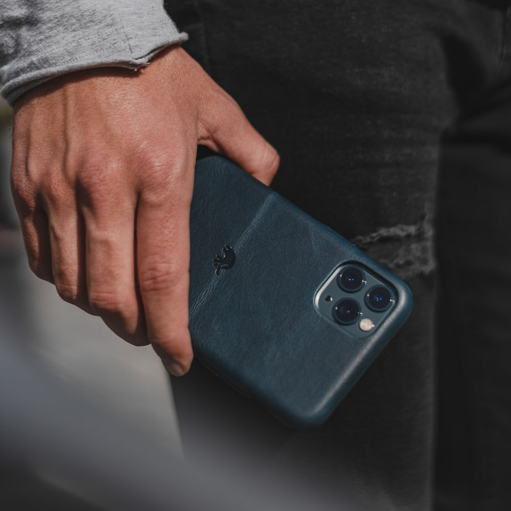 Hand holding dark ocean-blue leather iPhone case with embossed logo and triple camera cutout.