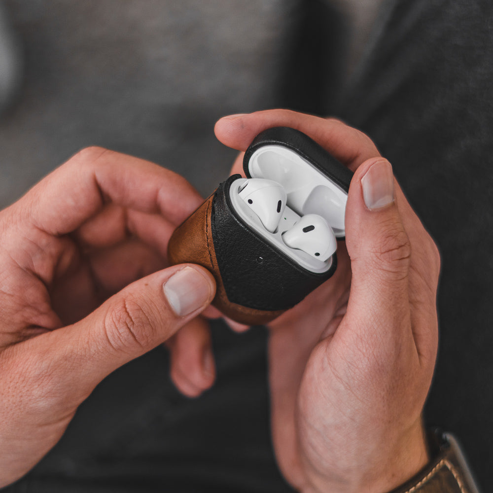 Brown-and-black leather AirPods case held open in hands, showing white AirPods inside (TERRA).