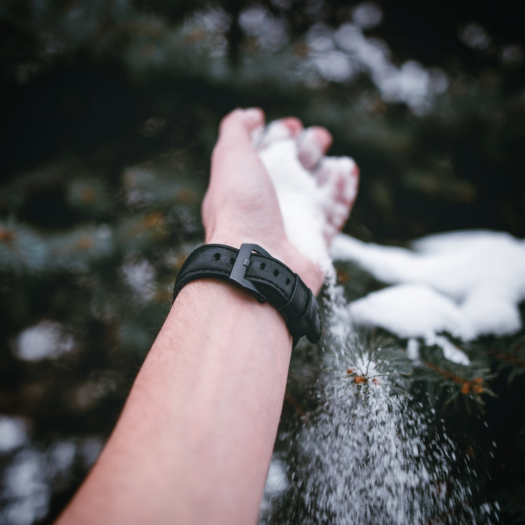 A hand wearing a matte black leather Apple Watch strap, with snow falling around it.