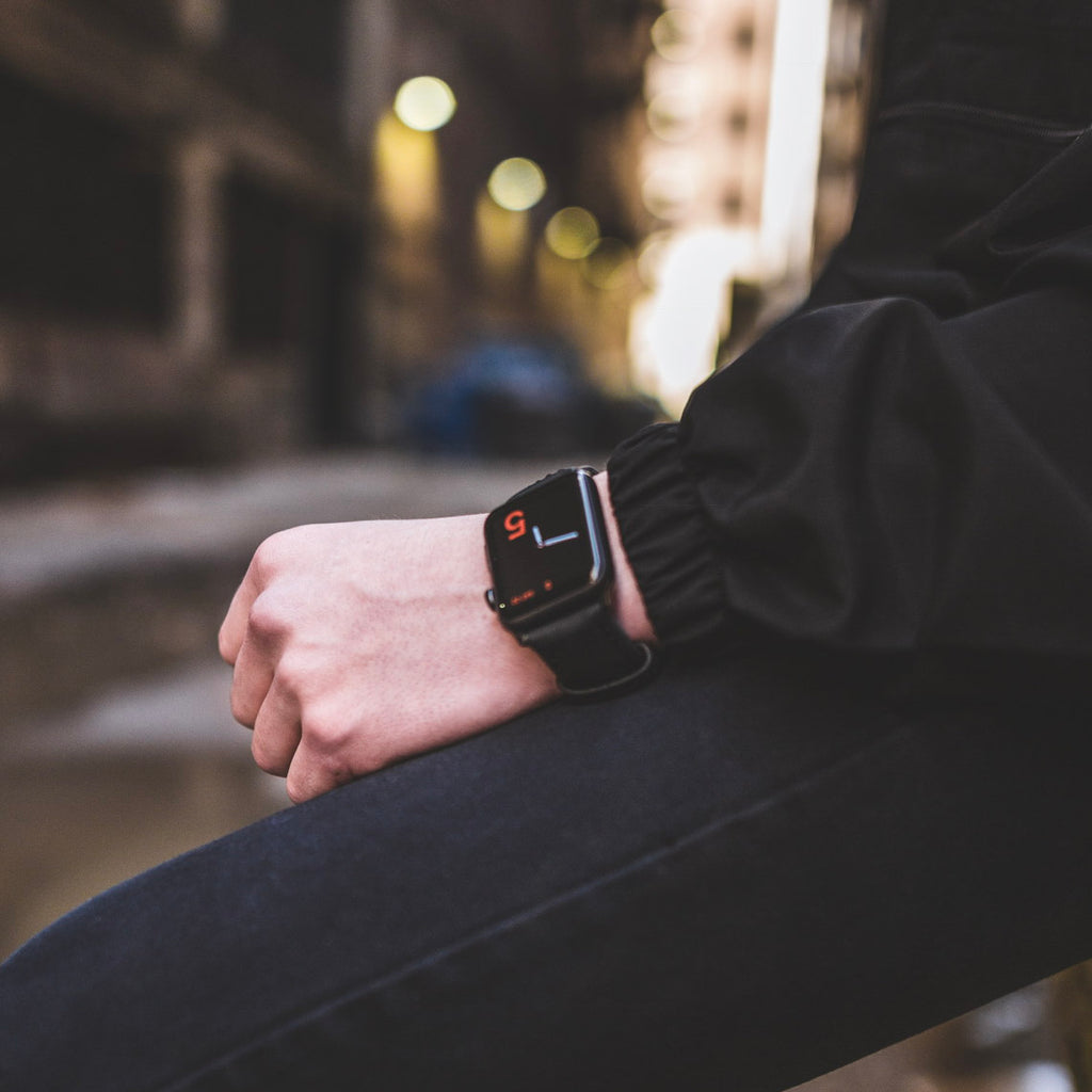 A person wearing a matte black leather Apple Watch strap on their wrist, with a blurred urban background.