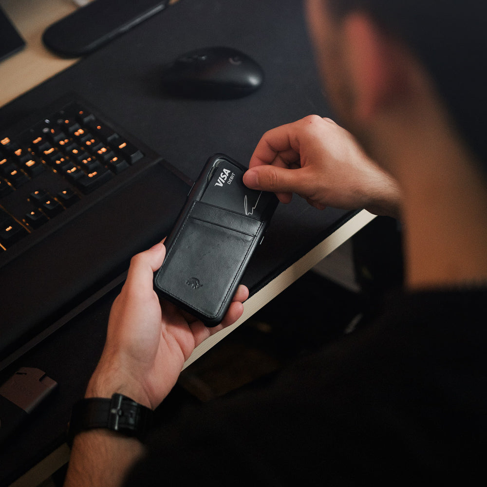 Hands sliding a VISA DEBIT card into a black leather phone-wallet card slot on a desk near a keyboard.