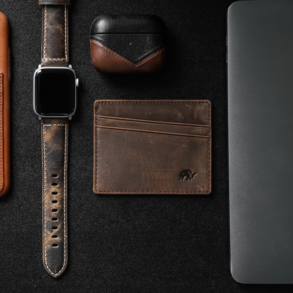 A brown leather card holder with four slots, placed next to a watch, AirPods case, and laptop.