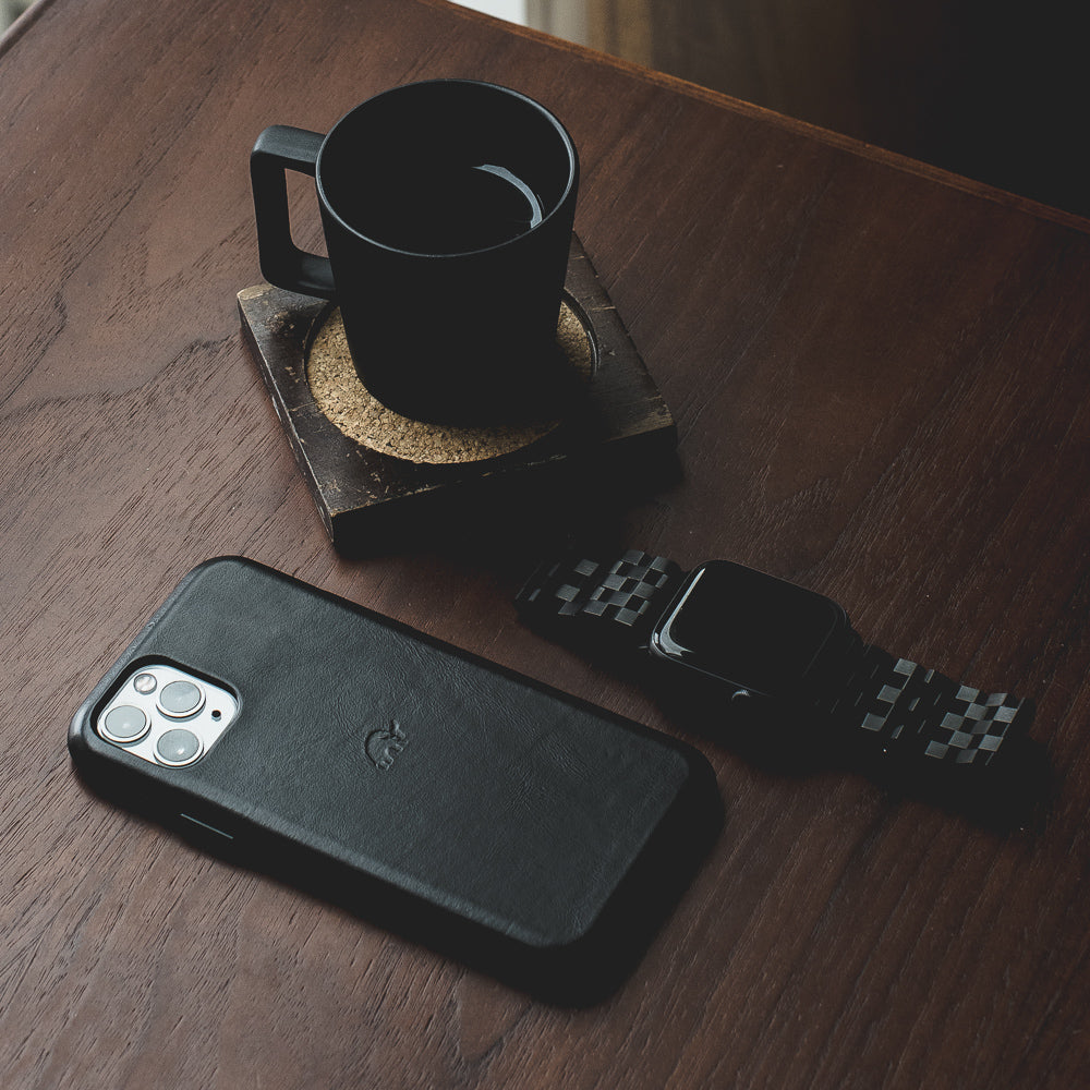 Black leather iPhone case on wooden table beside a black coffee mug on a cork coaster and a black smartwatch.