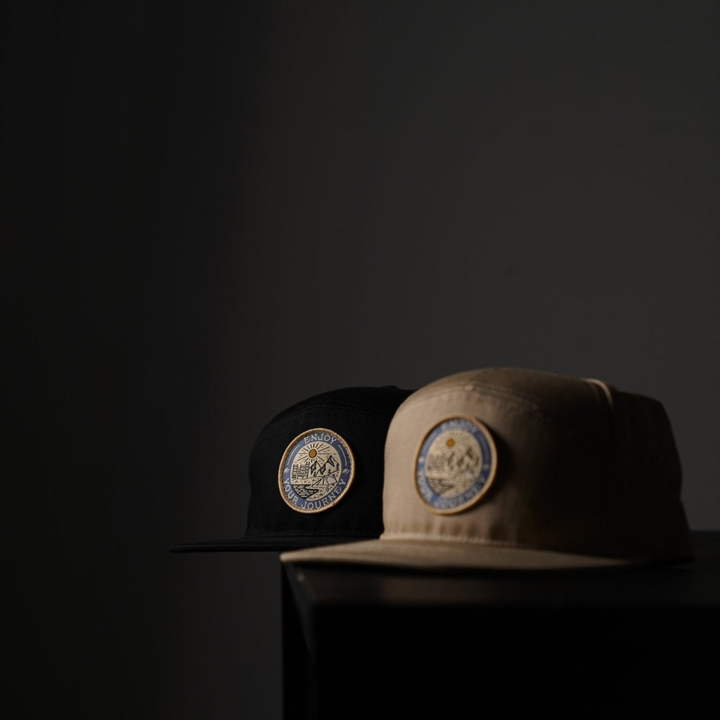 Black and tan caps with circular embroidered patch of sun and mountains reading 'ENJOY YOUR JOURNEY'.