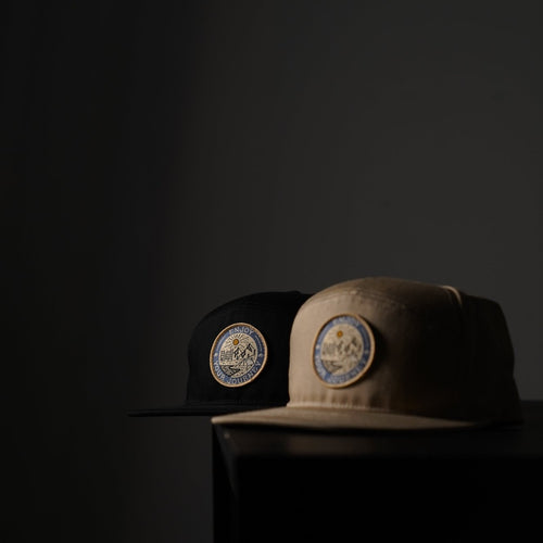 Black and tan caps with circular embroidered patch of sun and mountains reading 'ENJOY YOUR JOURNEY'.
