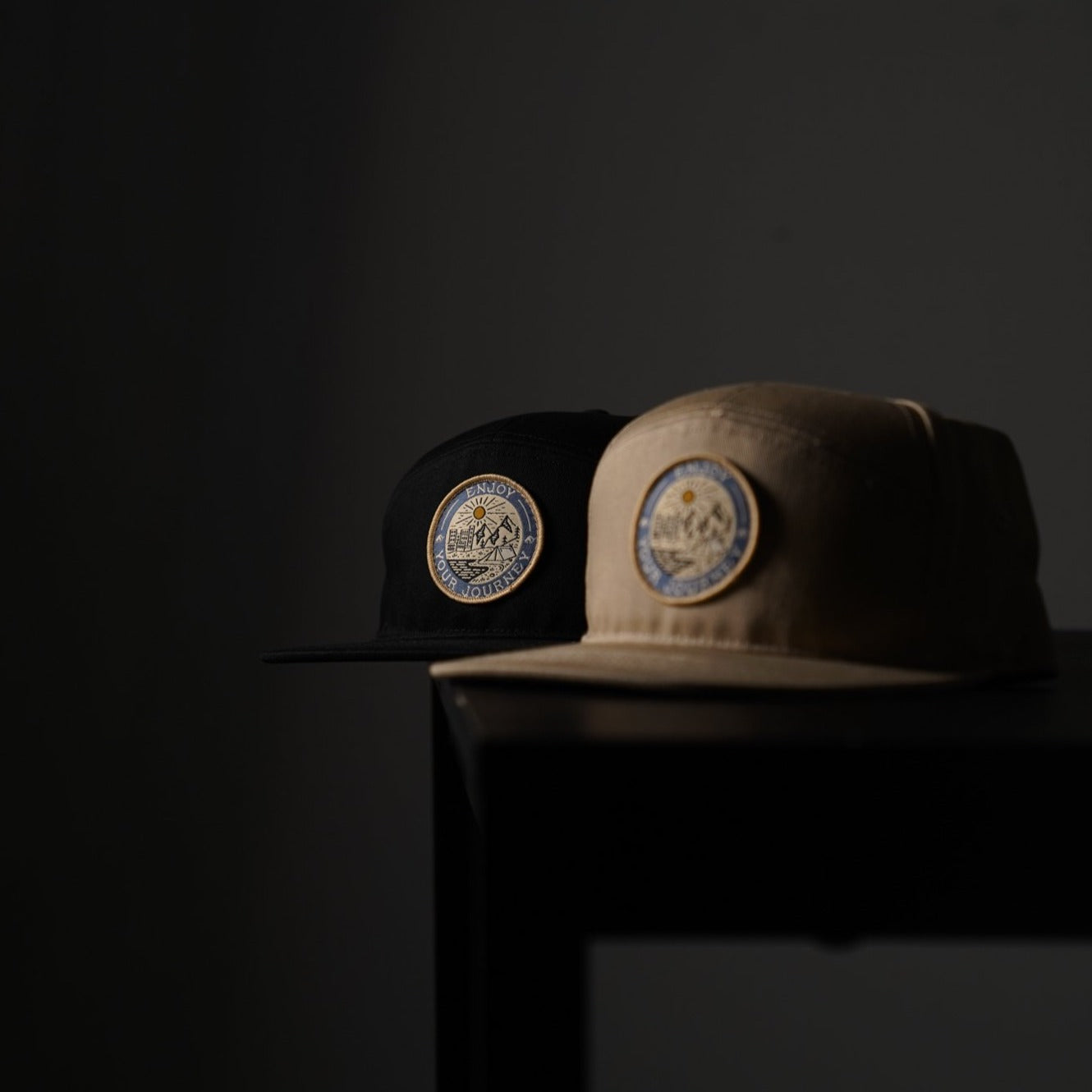 Two five-panel caps (black and tan) on a table, each featuring a round patch reading: ENJOY YOUR JOURNEY