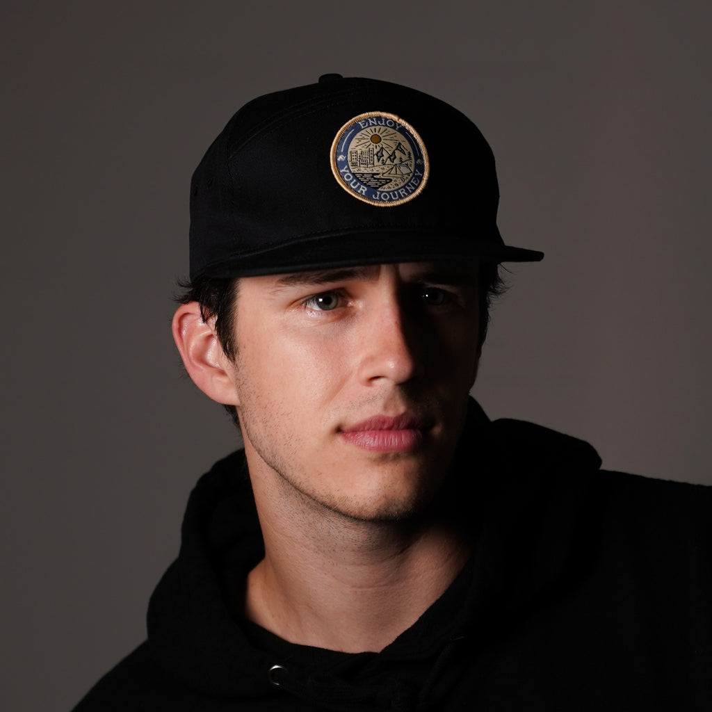 Black cap with round embroidered badge reading 'ENJOY YOUR JOURNEY', modeled by young man in black hoodie.