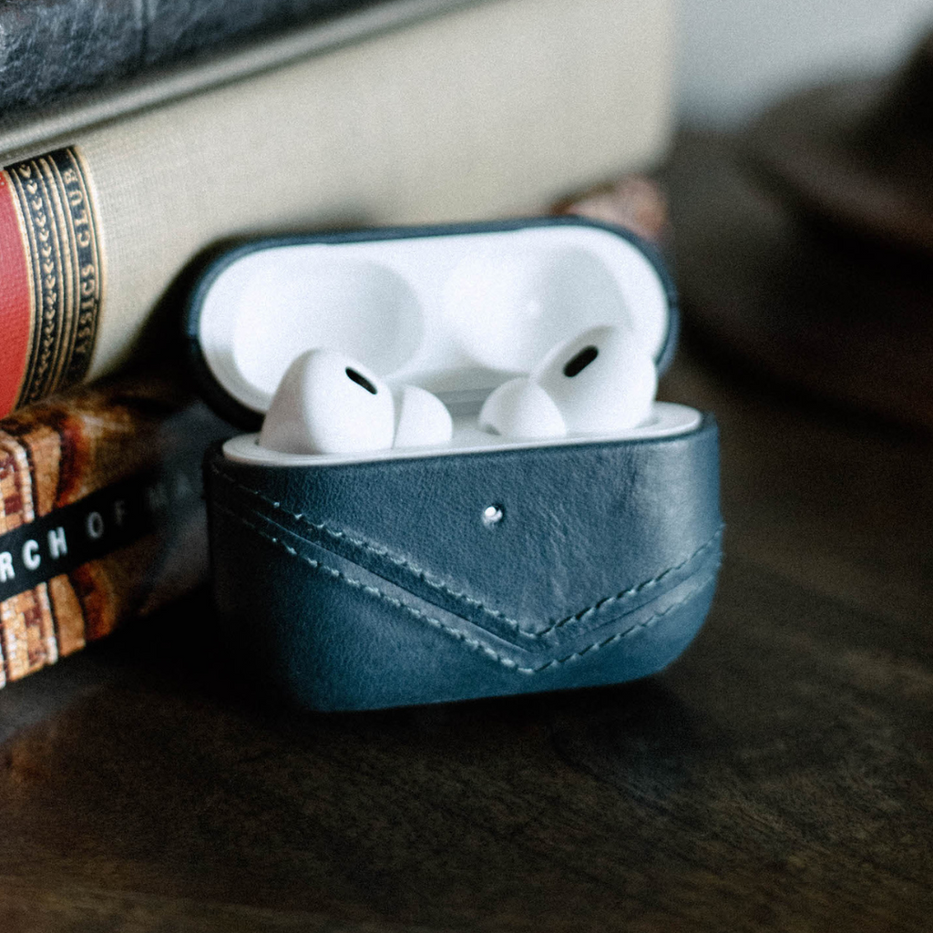 Leather AirPods case in Ocean color with AirPods inside, placed on a wooden surface next to books.