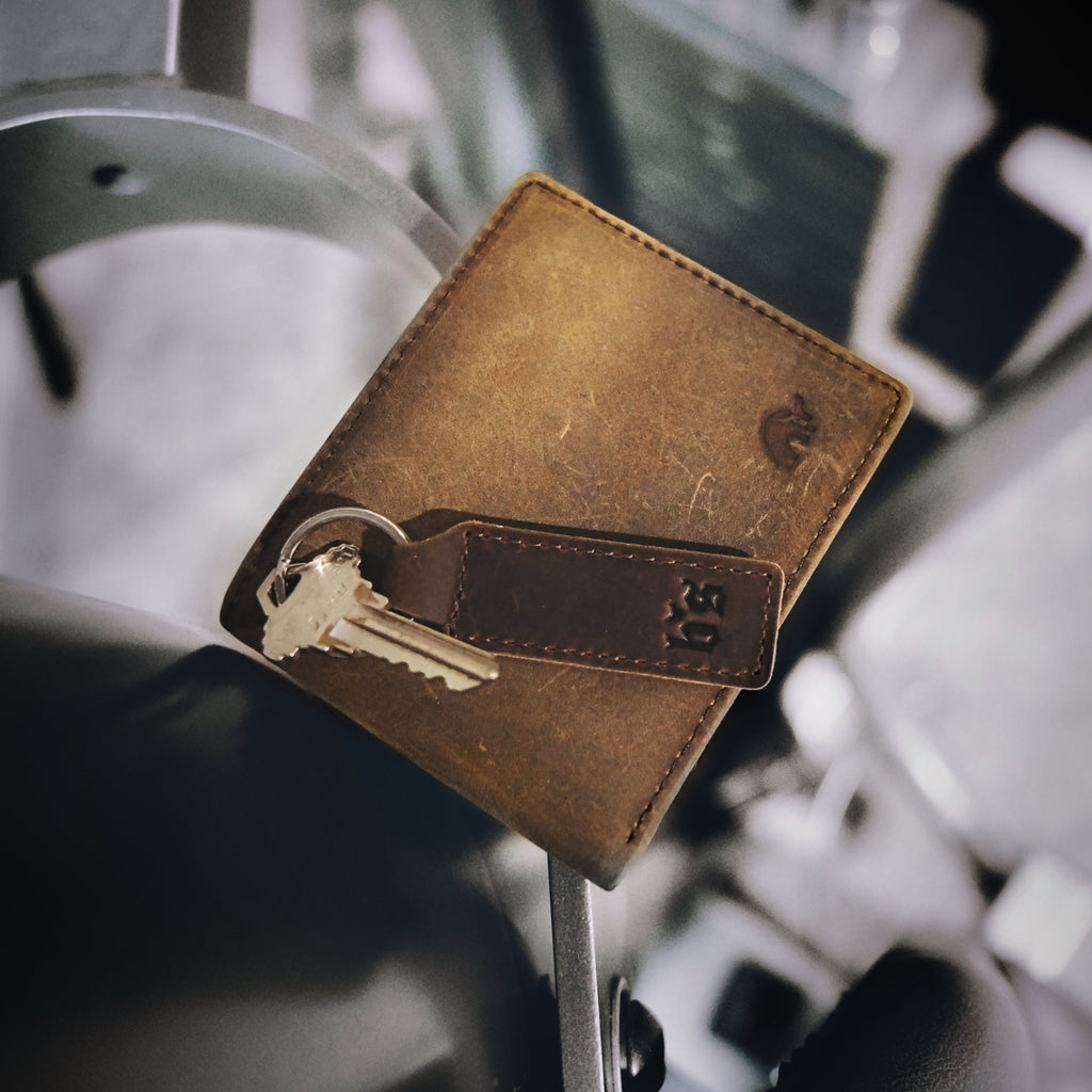 Worn brown leather bifold wallet with stitched flap, small embossed logo and attached keyring holding two keys