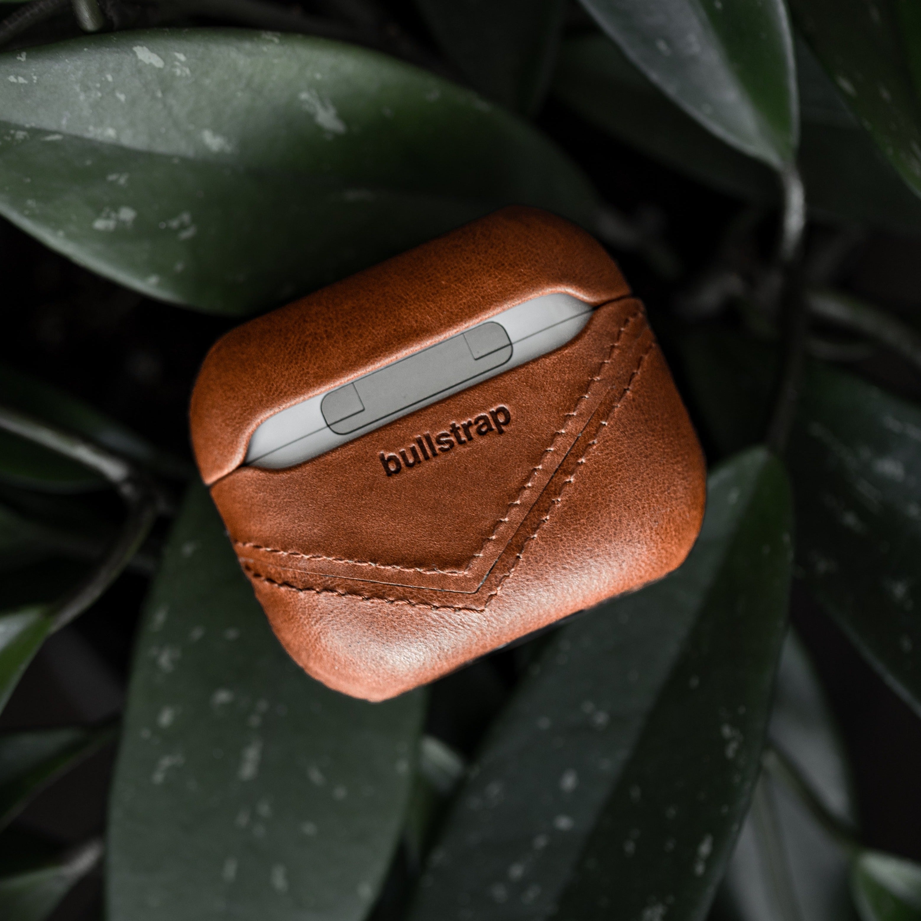 Leather AirPods case in sienna color with embossed bullstrap logo on a plant background