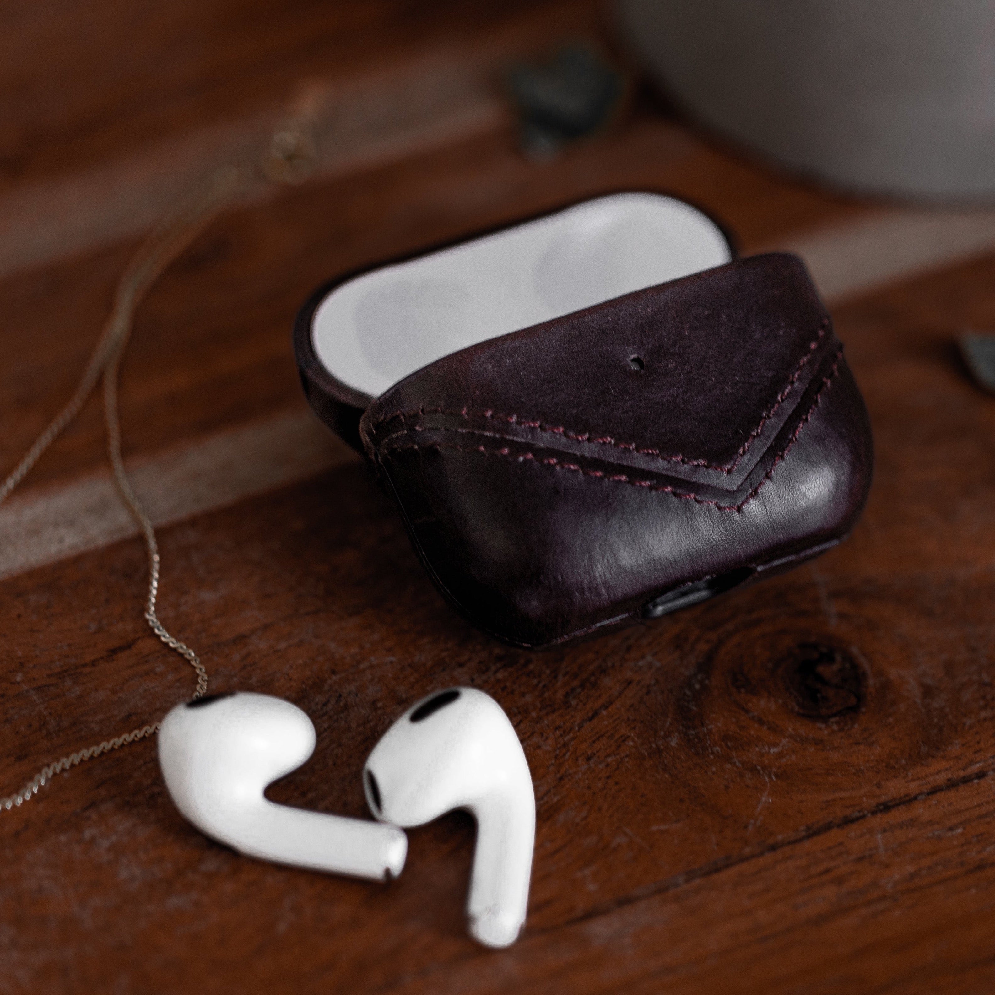 Leather AirPods case in bourbon color with AirPods beside it on a wooden surface.
