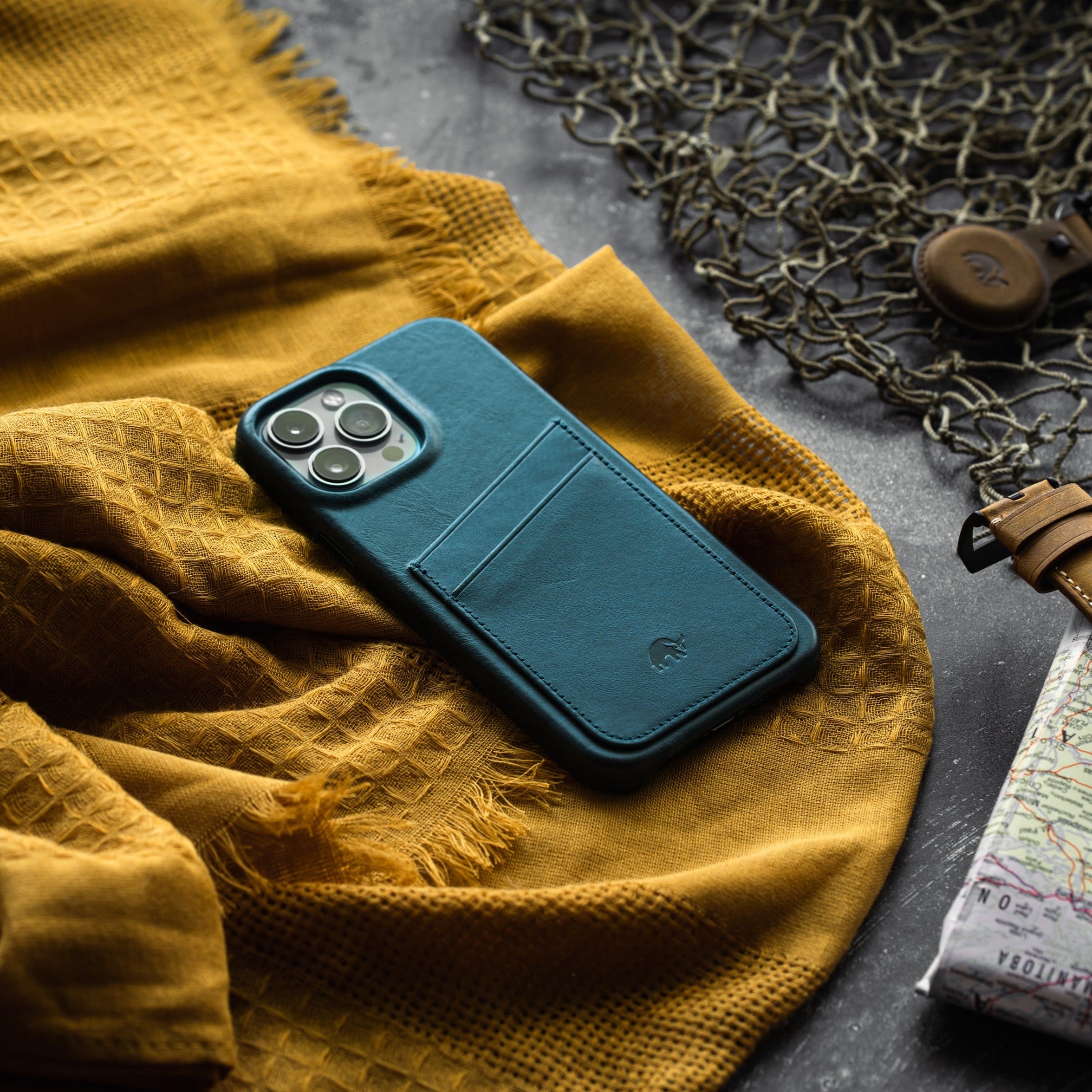 Teal leather phone wallet case with two-card back pocket on mustard textured cloth next to map and leather strap