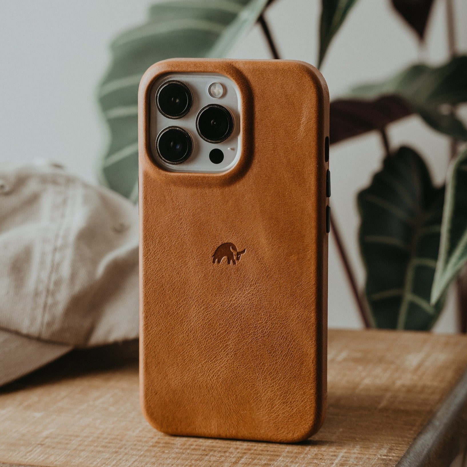 Sienna brown leather phone case with raised camera cutout and embossed elephant logo, standing on a wooden table
