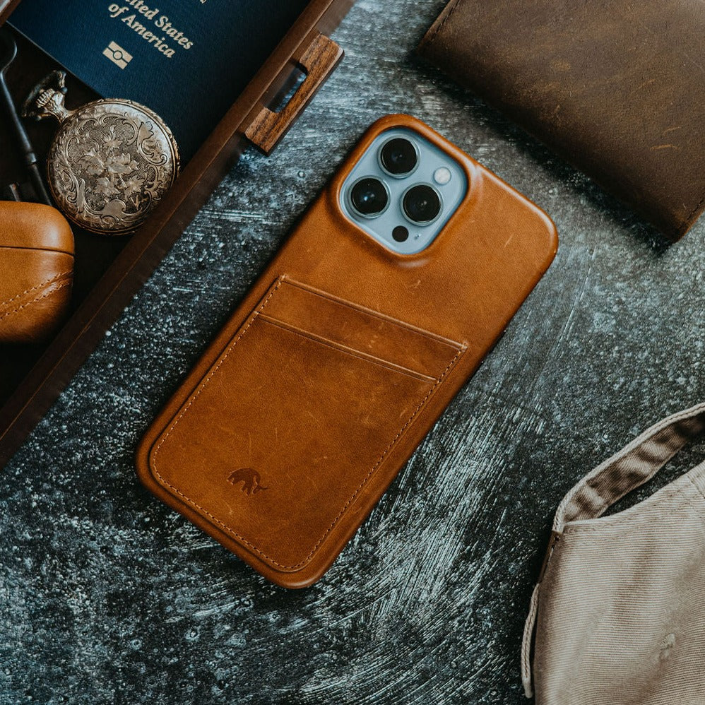Leather brown wallet-style phone case on tabletop with passport reading 'United States of America', pocket watch and canvas bag.