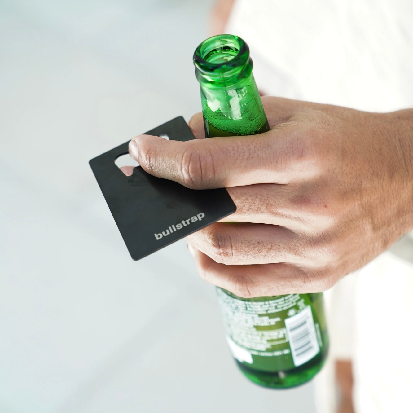 A person holding a bottle with the Bullstrap Bottle Opener Card in hand, featuring the 'bullstrap' logo.
