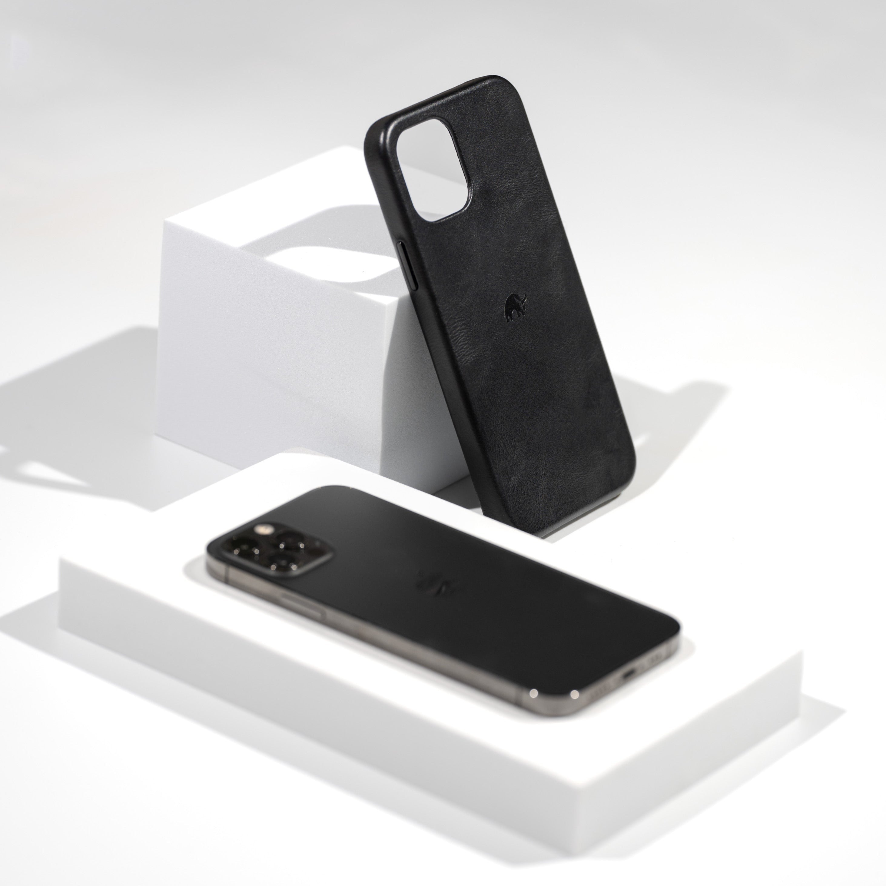 Black leather minimalist phone case standing on a white pedestal beside a black smartphone