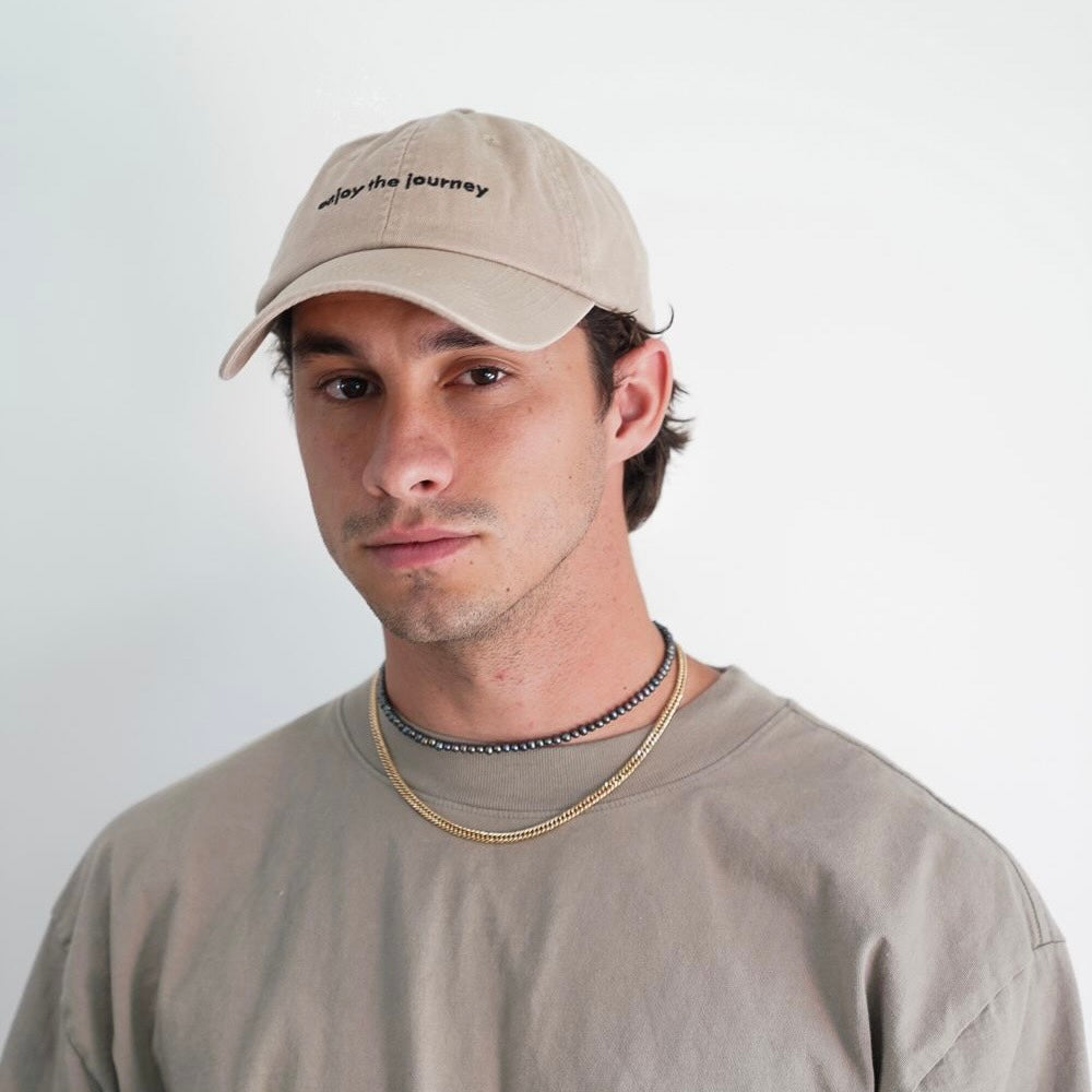 Man wearing tan dad hat embroidered 'enjoy the journey' with tan shirt on neutral background