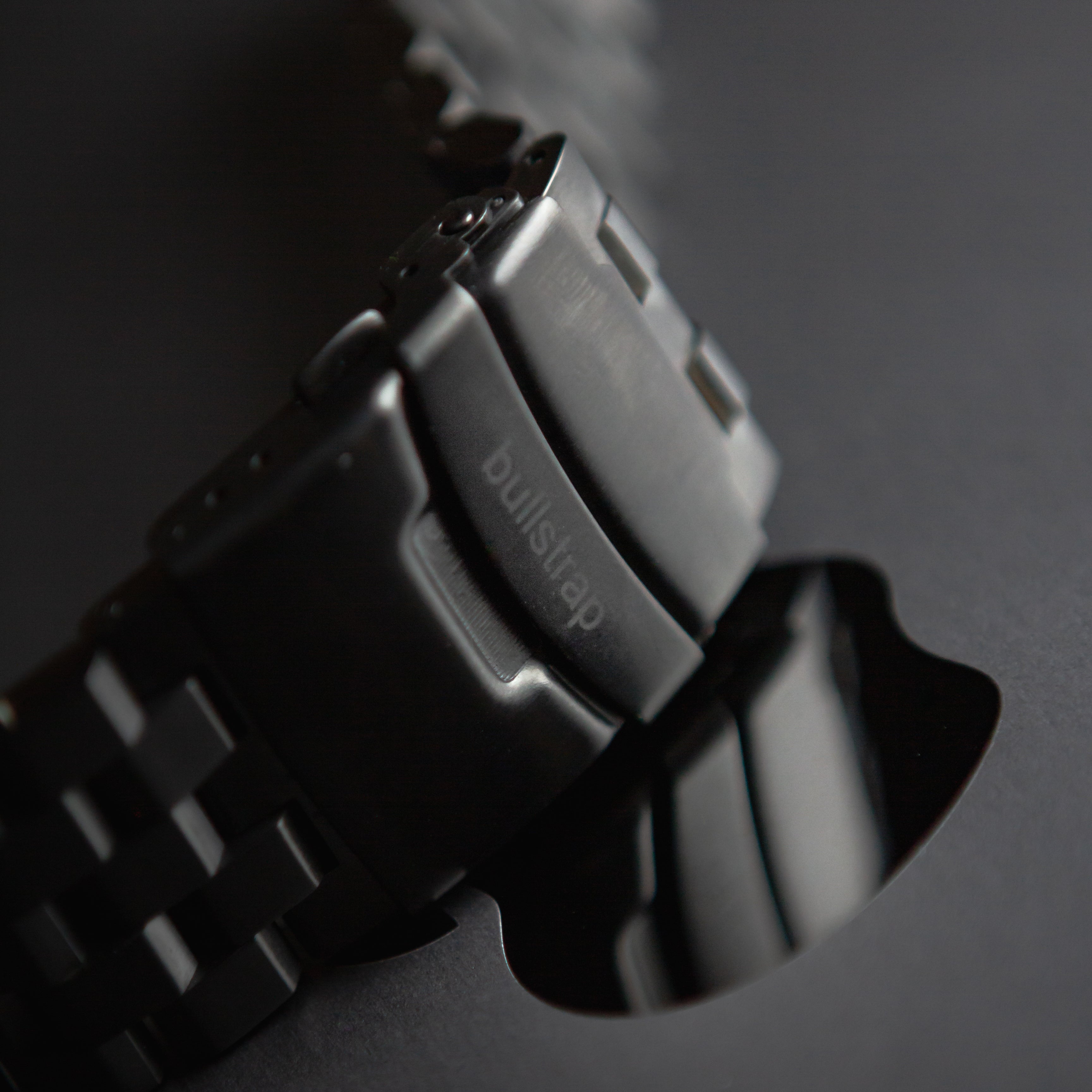Black metal link Apple Watch strap, close-up of clasp engraved 'bullstrap'