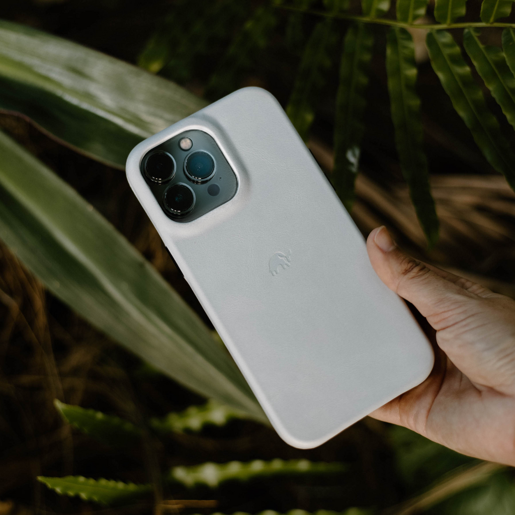 White minimalist iPhone case with embossed elephant logo, held in a hand over green foliage.