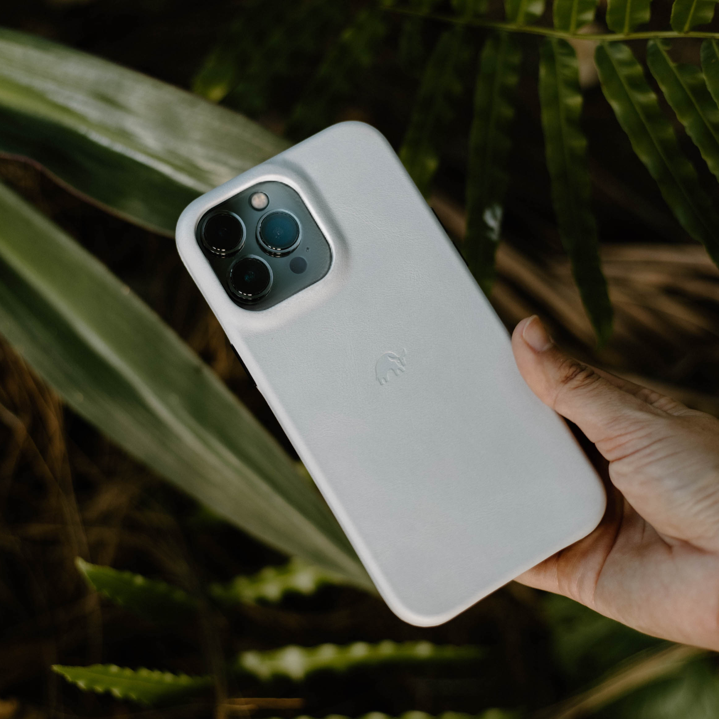 White minimalist iPhone case with embossed elephant logo, held in a hand over green foliage.