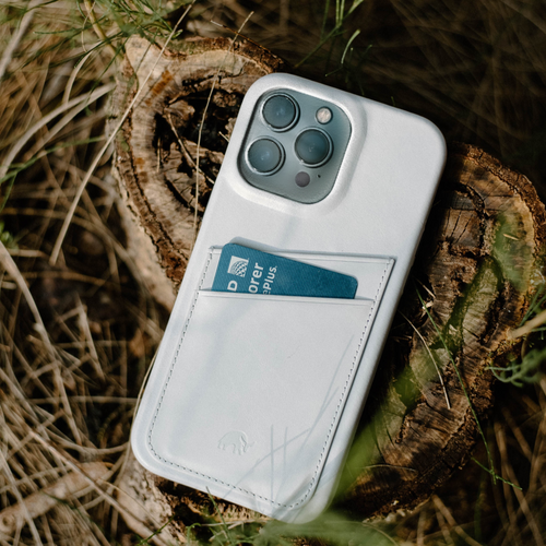 White leather iPhone case with card pocket holding a blue card reading "Explorer ePLUS" on a tree stump