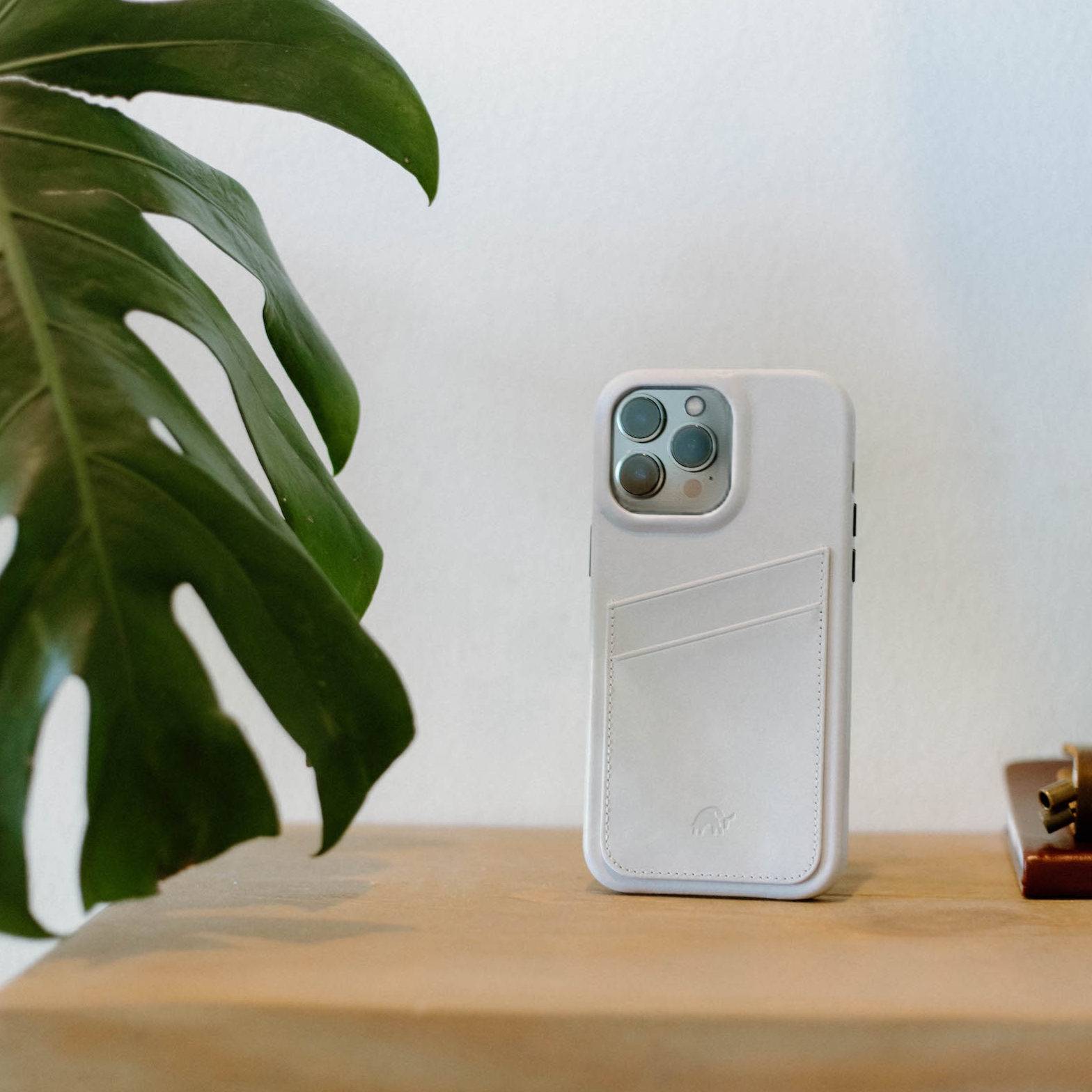 White leather iPhone case with stitched card pocket standing on a wooden table beside a large green plant leaf.